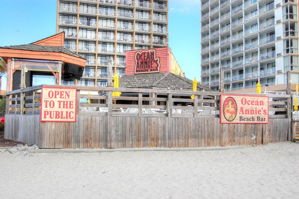 A lively beach bar scene featuring the Ocean Annie's Beach Bar signage, wooden fencing, and the beach setting in the background. Guests are seen socializing, enjoying drinks, and listening to live music as the sun sets over the ocean, creating a festive and relaxed atmosphere.