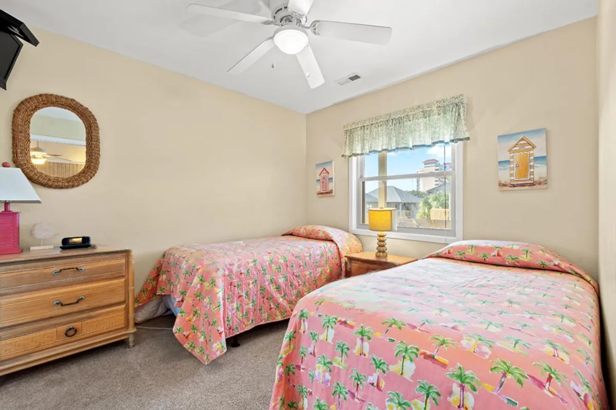 A light-filled twin bedroom with two beds decorated with floral bedding, a nightstand with a lamp between them, a decorative mirror, and a window offering views outside. The space feels welcoming and practical for families or groups.
