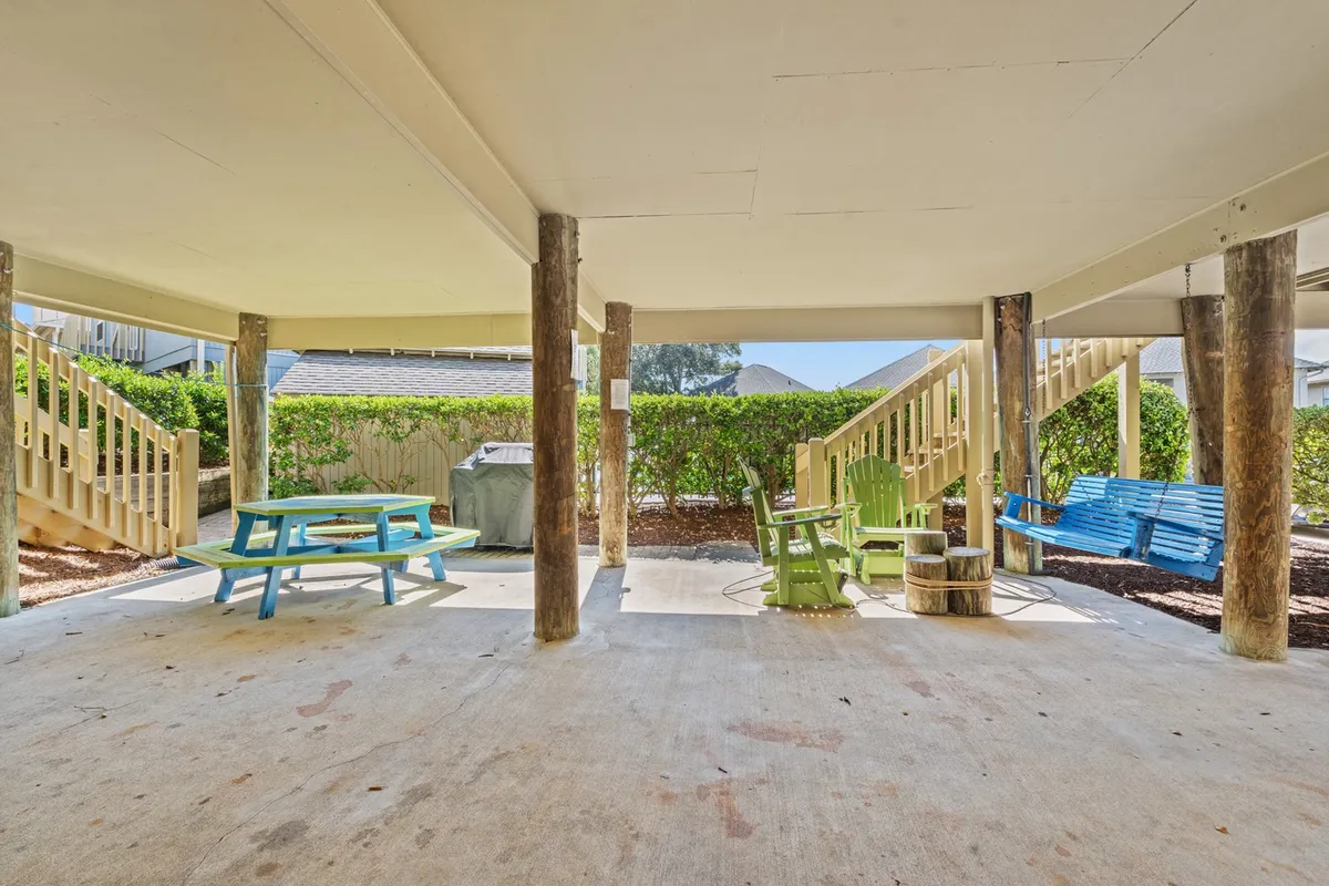 This exterior shot features a colorful picnic table, green Adirondack chairs, a blue bench, and sturdy wooden posts beneath the elevated cottage. The area is shaded, creating a comfortable space for relaxing and social gatherings. The under-house setup clearly displays the covered parking area as well as an informal, resort-like outdoor lounge zone.