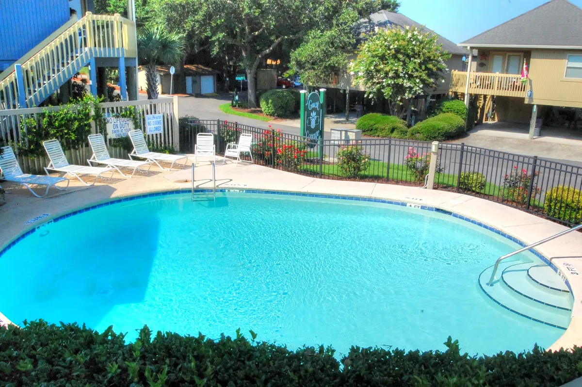 The image showcases the inviting exterior of a spacious cottage in Myrtle Beach, with a sparkling swimming pool surrounded by comfortable lounge chairs, lush landscaped gardens, and a staircase leading up to the private entrance. The setting is perfect for relaxation and social gatherings, hinting at sunset swims, poolside barbecues, and tranquil mornings.