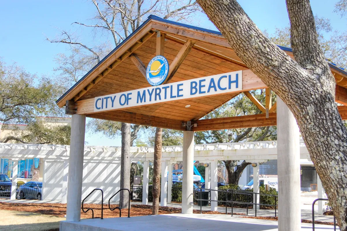 This image features the welcoming signage of 'CITY OF MYRTLE BEACH' on a wooden structure surrounded by lush greenery and trees. The scene suggests a gathering place for visitors seeking information or joining local community events, setting the tone for vacation exploration and local adventure.