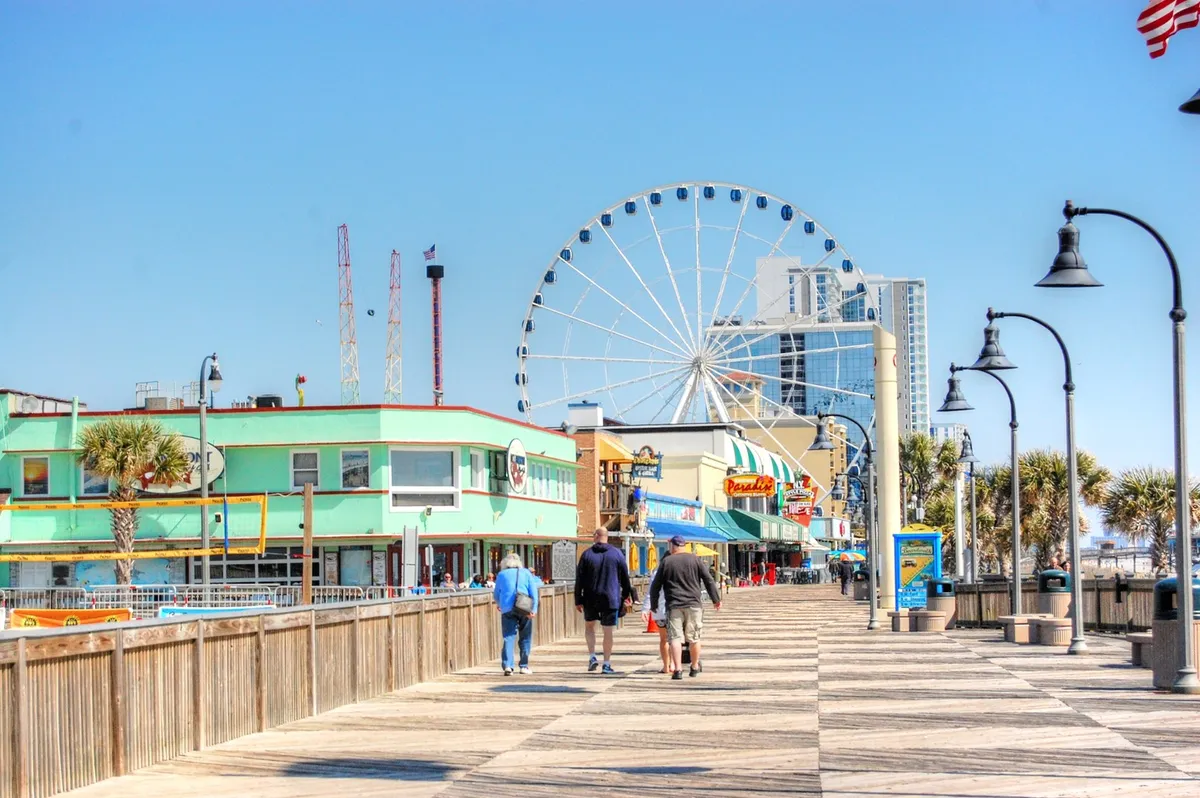 A bustling coastal boardwalk scene featuring a towering ferris wheel (SkyWheel), colorful waterfront buildings, and families strolling, shopping, and enjoying various rides at sunset. People can be seen savoring ice cream and soaking in the joyful atmosphere.