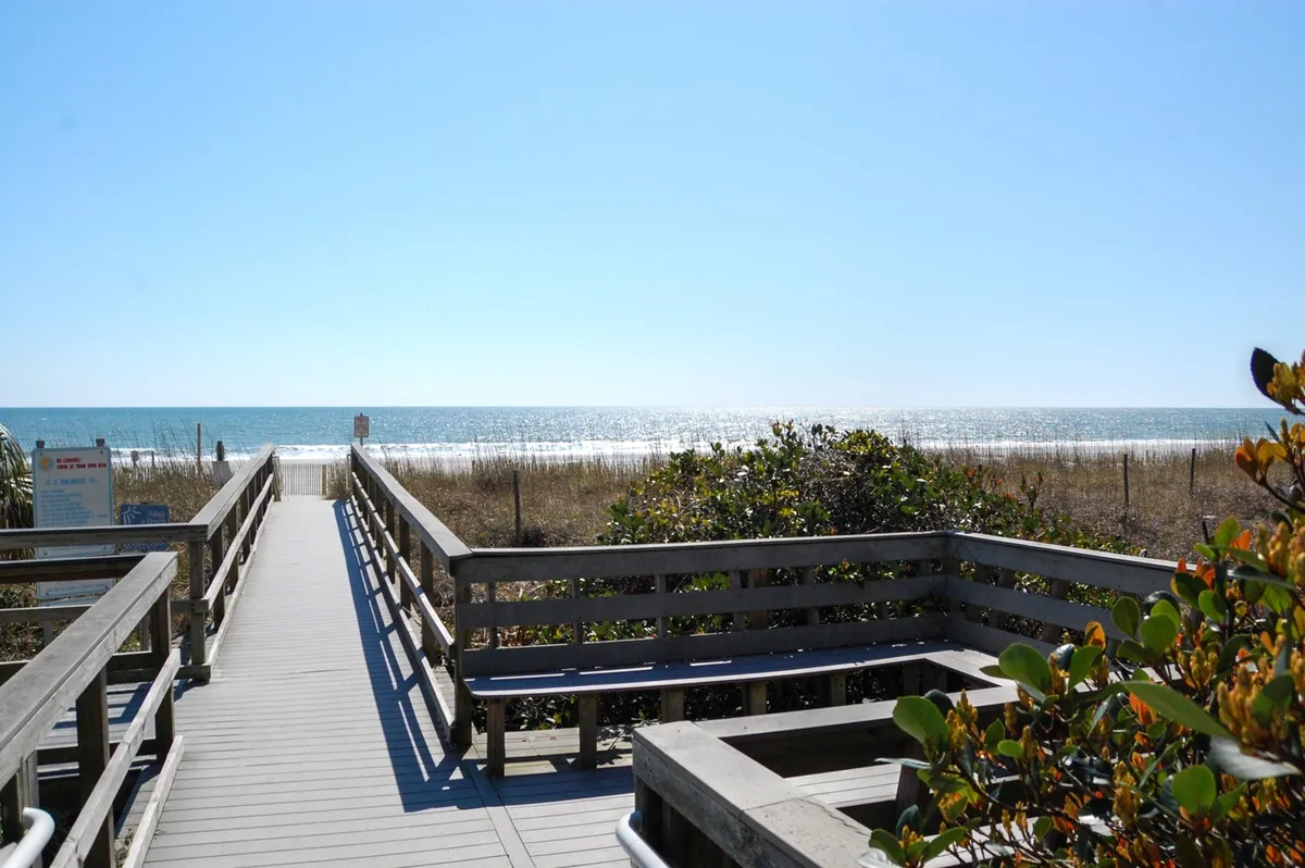 A wooden boardwalk leads over dune grasses toward an expansive ocean view, with sunlight illuminating the path and hints of sandy beach visible. This scene highlights the close proximity and easy access from the property to Myrtle Beach’s famous shoreline.