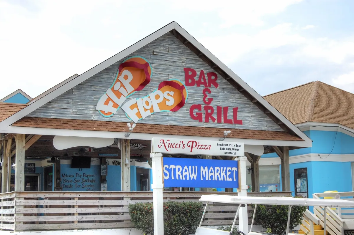 A bustling exterior with visible signage for Flip Flops Bar & Grill, Nucci's Pizza, and the Straw Market. The scene is alive with people dining, shopping, and enjoying live music events, capturing the spirited, social ambiance of popular Myrtle Beach hangouts.